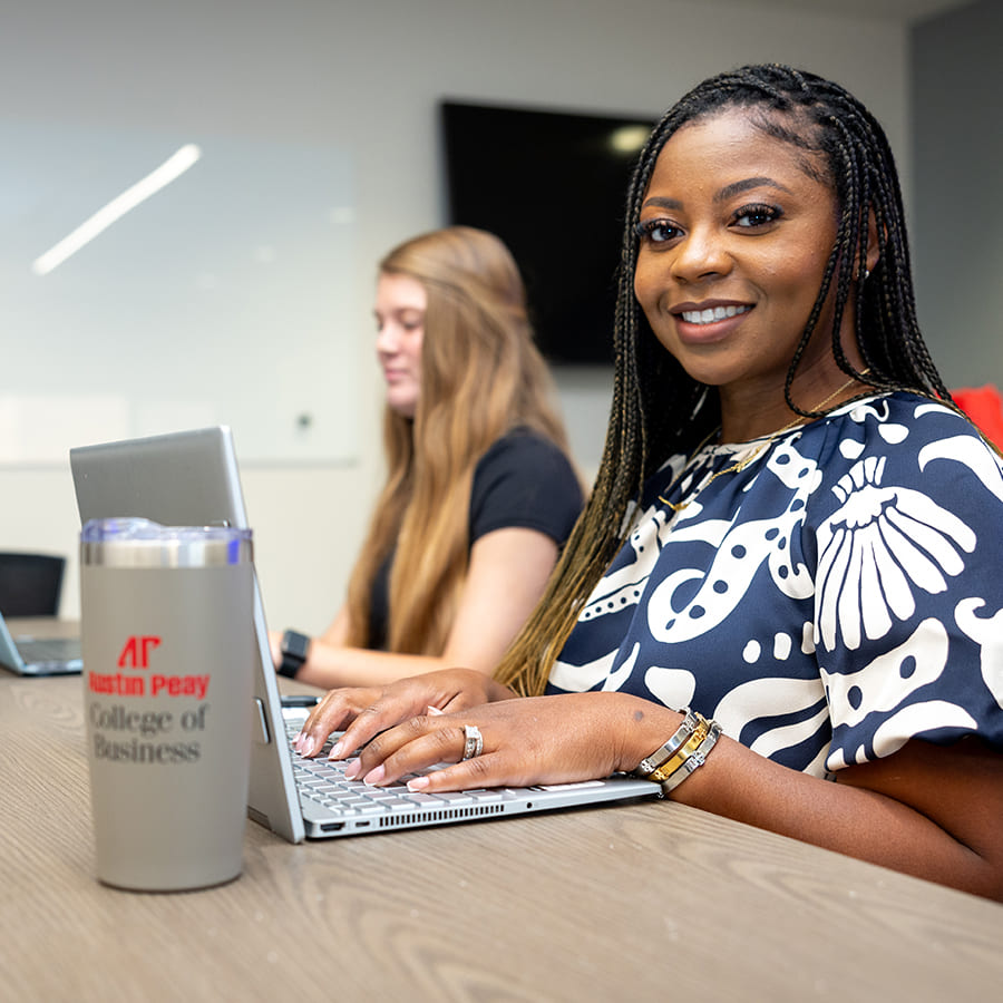 Students sitting at a conference table working on laptops with the one in the foreground smiling at the camera.
