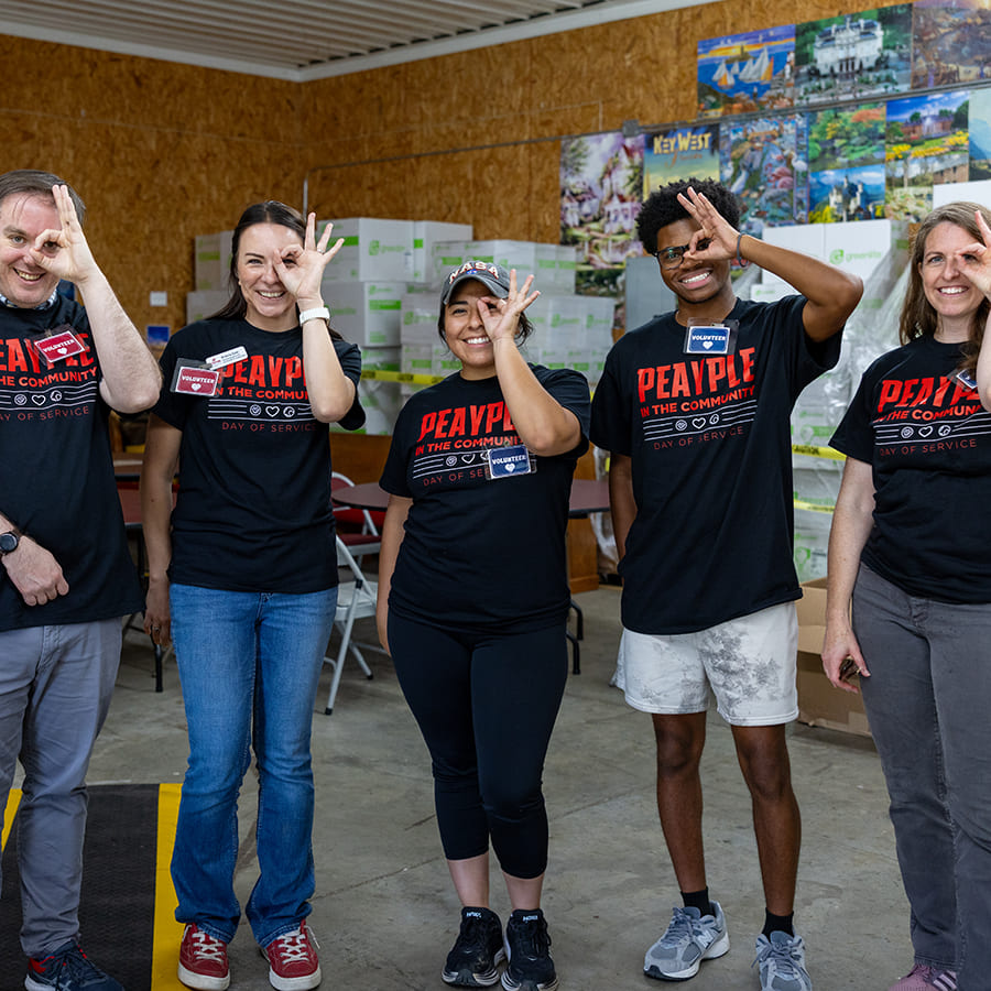 Group of Students and Employees wearing black 'Peayple in the Community Day' t-shirts standing in a line and holding up the monocle ('ok') hand sign over their left eyes.