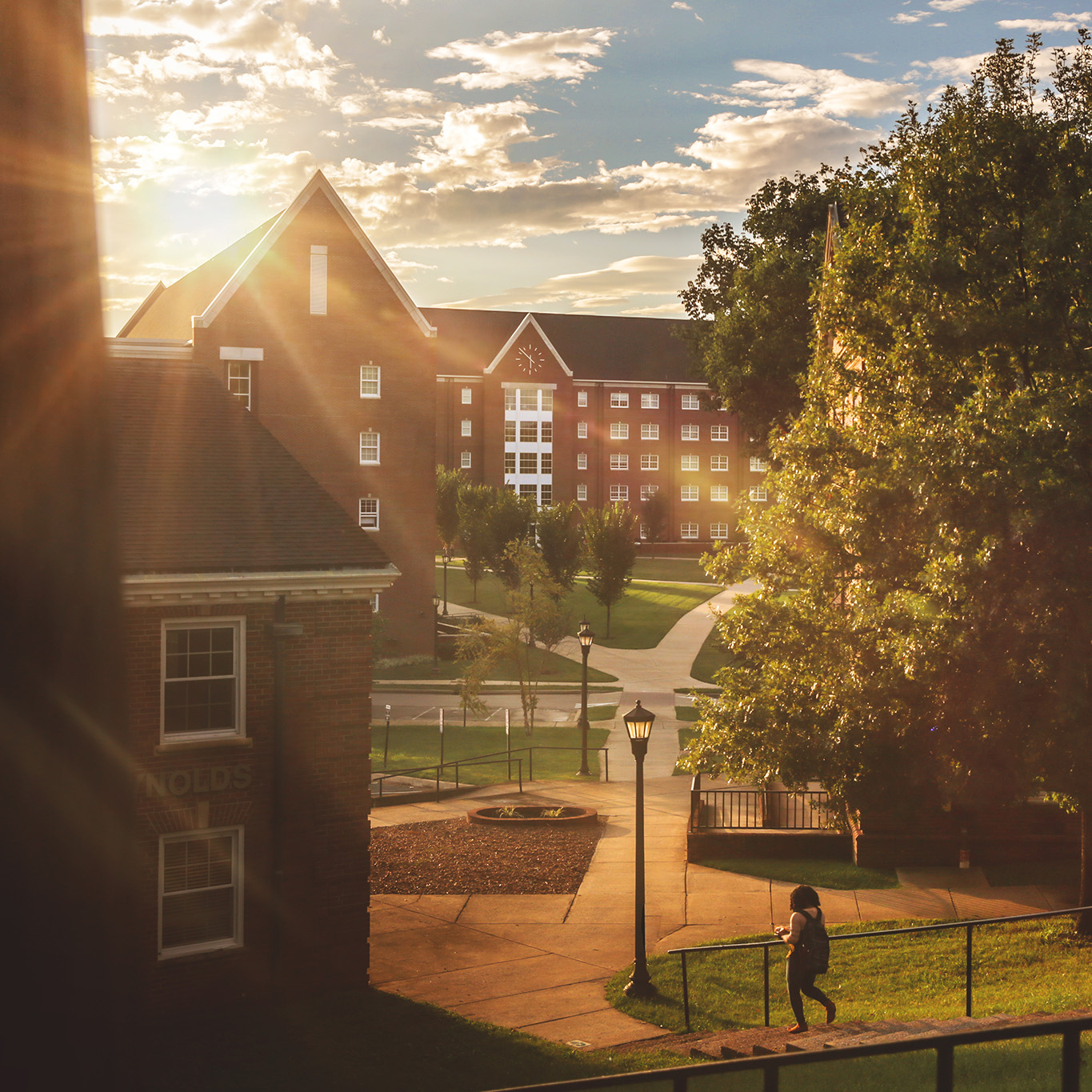 Sunset, golden hour, over red-brick campus buildings, walkways, and trees, with a person walking down a set of stairs.