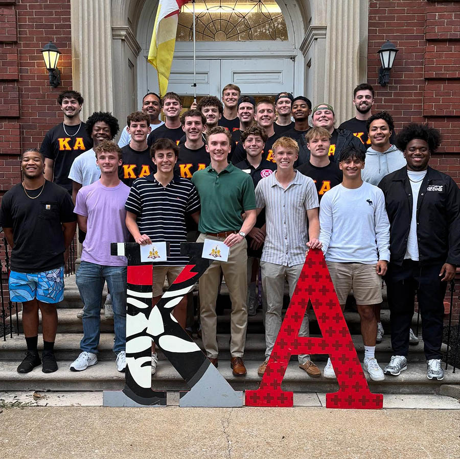 Members of the Kappa Alpha fraternity standing on the stairs of the KA house with giant 'KA' letters standing on the ground in front of them.