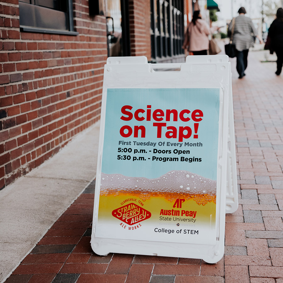 An a-frame sign on the sidewalk next to a brick building that reads 'Science on Tap' with people walking away in the distance.