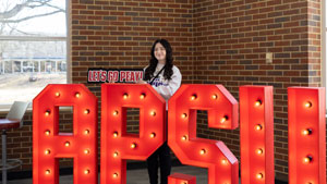 Student holding a Let's Go Peay! sign, while standing behind giant light up letters that spell APSU.