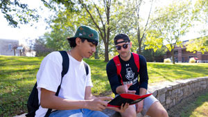 One student showing another something in their notebook while they sit outside.