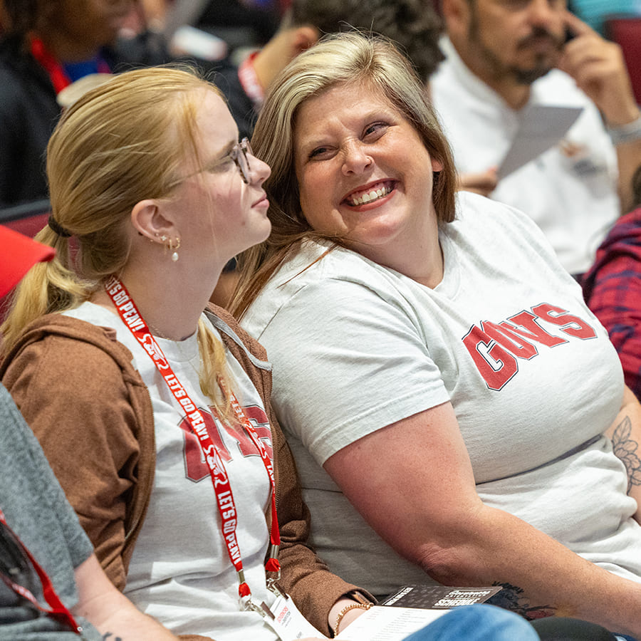 A smiling mother and daughter, wearing matching "GOVS" shirts, share a joyful moment together while sitting in the audience for new student orientation.